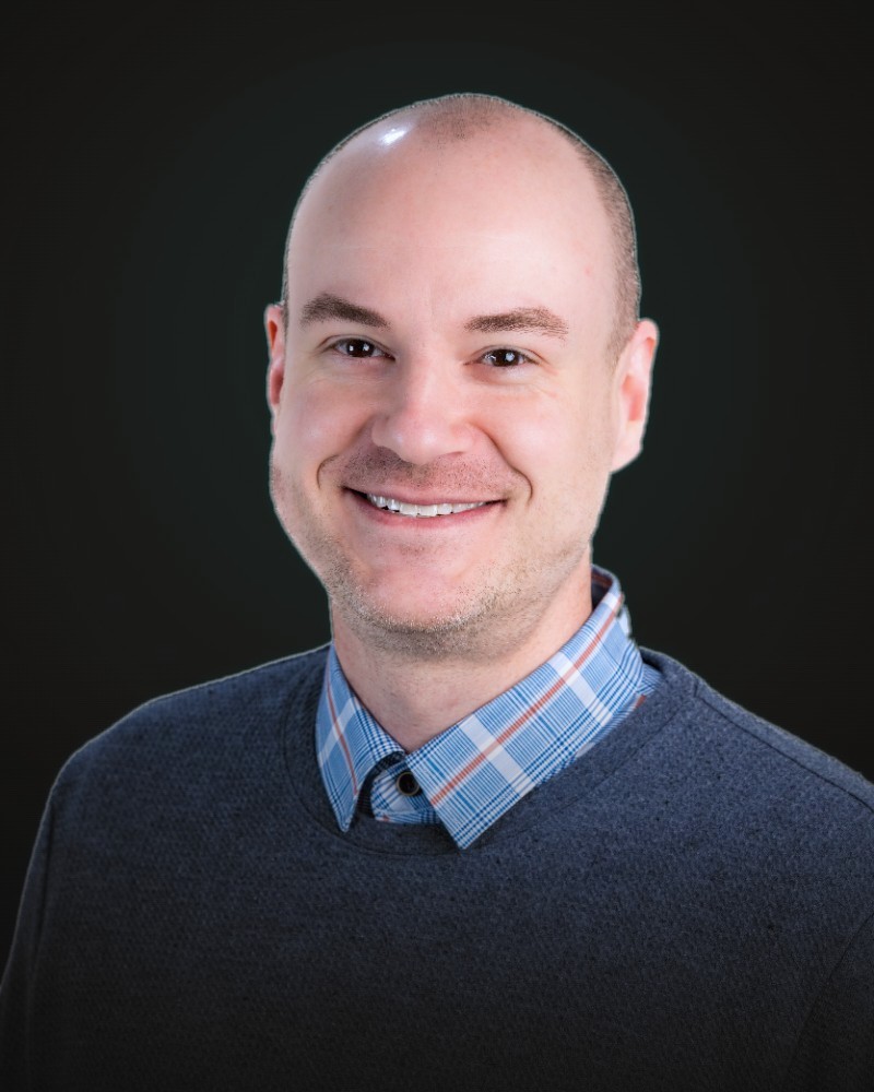 Portrait of John Foster, smiling, against a dark studio background.